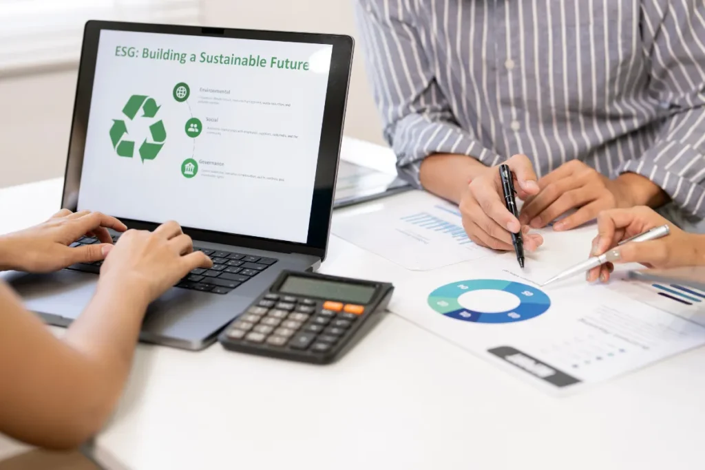 Profissionais analisando relatório ESG em notebook com gráfico de sustentabilidade enquanto discutem dados financeiros na mesa